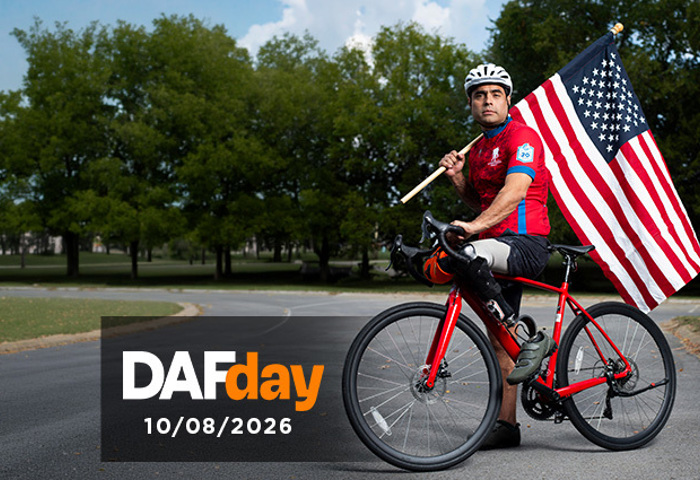 Warrior, Sal Gonzalez on a red bike with his prosthetic leg, holding a large American flag on a paved road with trees in the background.