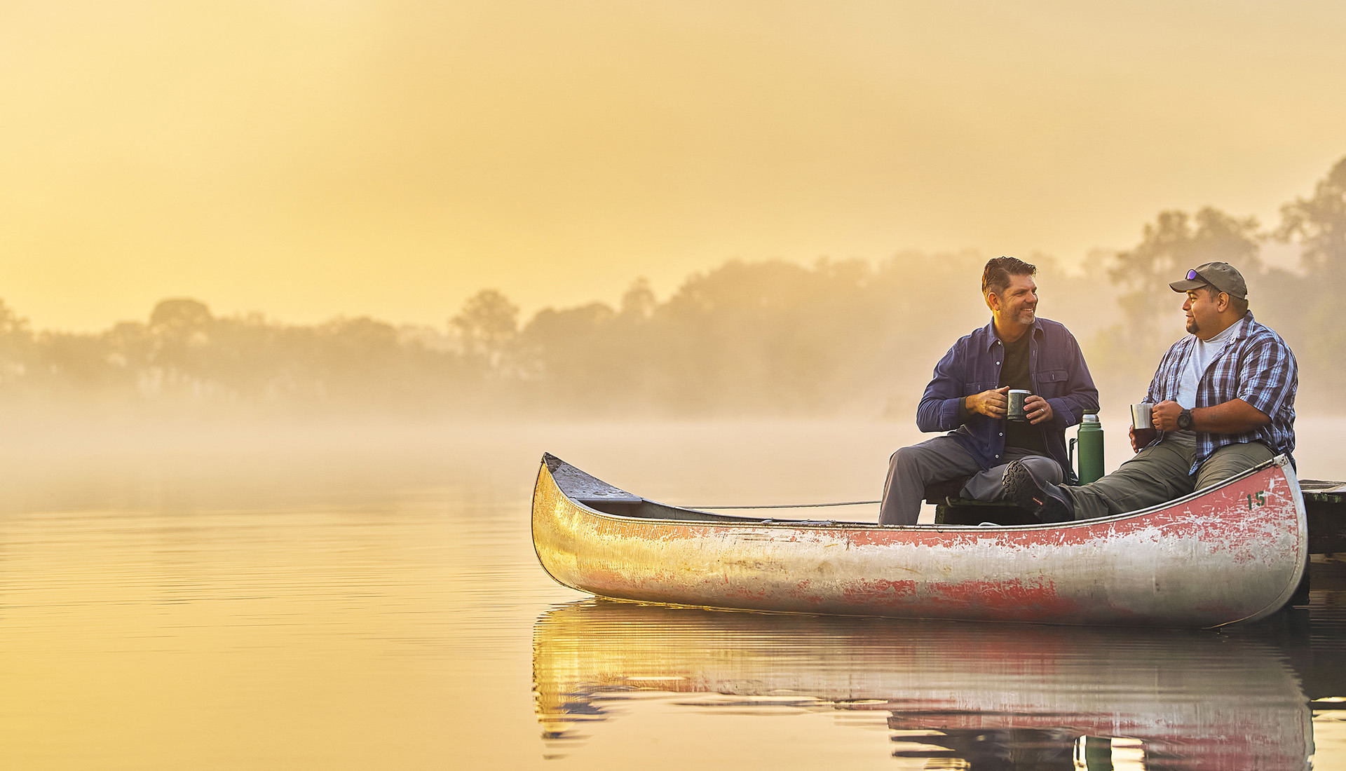 Warrior Tim Aponte and a fellow veteran sit at the edge of a dock at sunrise with their feet resting in a canoe.