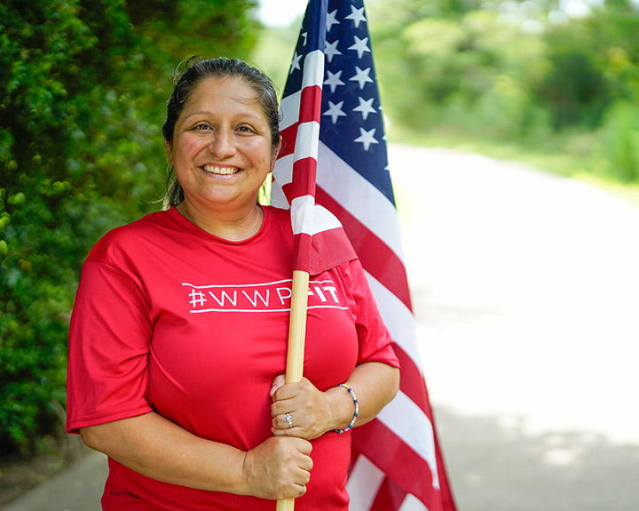 Wounded warrior Maria Edwards wears a red WWP Fit t-shirt while smiling and holding an American Flag along a tree-lined path.