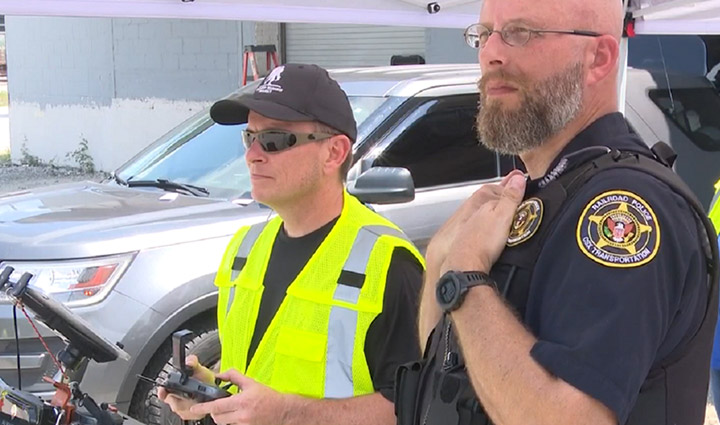 A wounded warrior wearing a yellow safety vest and sunglasses is operating the controls of a drone while a CSX police officer stands by him providing instruction.