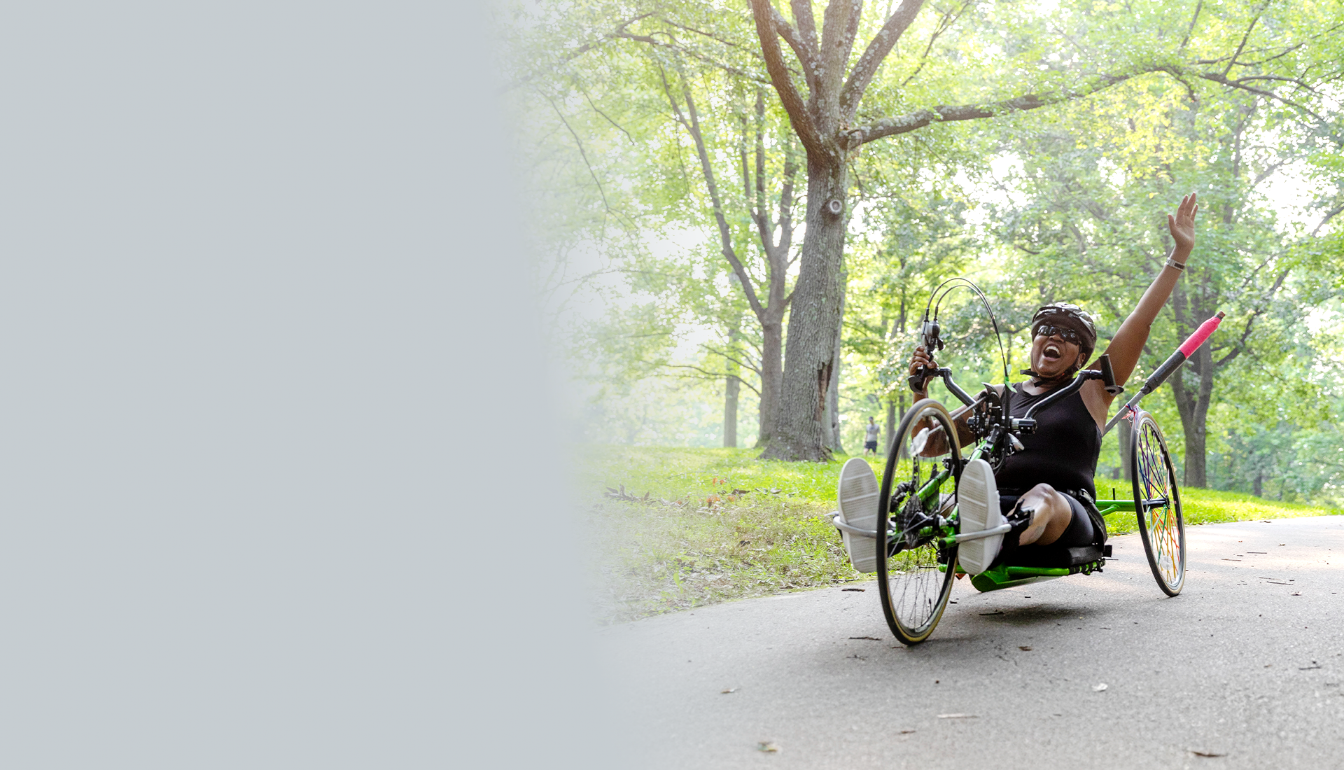 Wounded warrior Donna rides a recumbent bike on a path through a park, raising her arm in joy and accomplishment.
