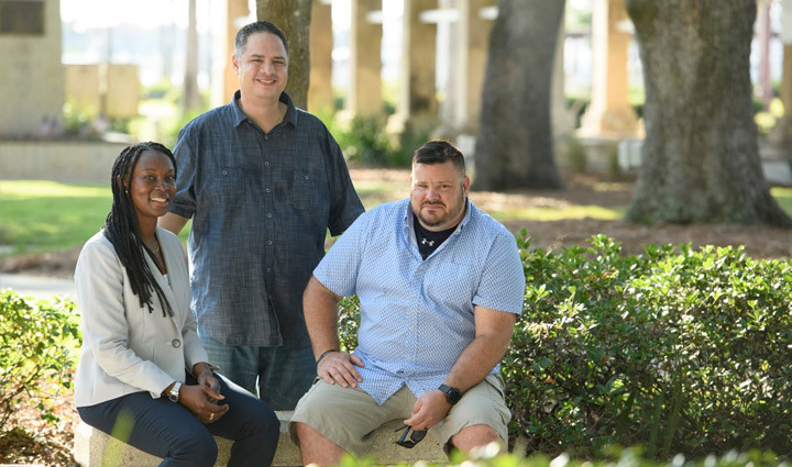 Wounded warrior Danielle Green sits on a bench outside next to wounded warrior Jake Norotsky with wounded warrior Jason Major standing behind them.