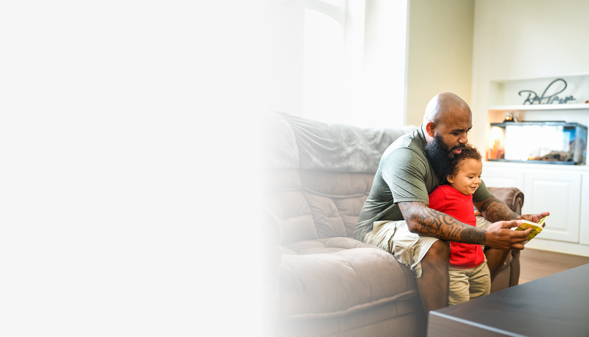 Warrior Eric Delion sits on a couch, wearing a green shirt and khaki shorts while reading to his son, who is wearing a red shirt and khaki shorts.
