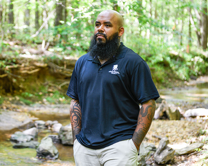 Wounded warrior Eric Delion stands in front of a stony brook surrounded by trees, appearing to be deep in thought while his hands are in his pockets.