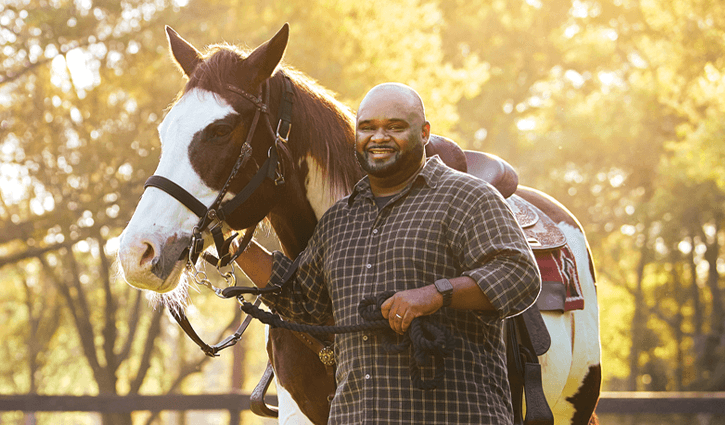 WWP warrior Chris Gordon smiles, holding the reins of a saddled brown and white horse on a sunlit ranch with trees and a fence.