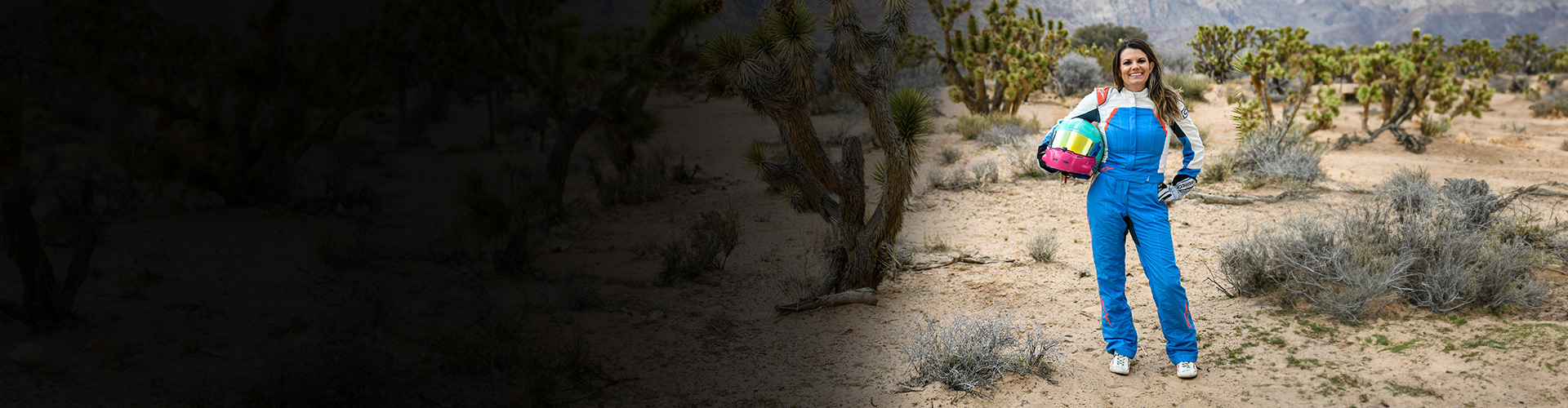 Wounded warrior Melissa McMahon stands in the desert wearing her race car jumpsuit and holding her helmet. 