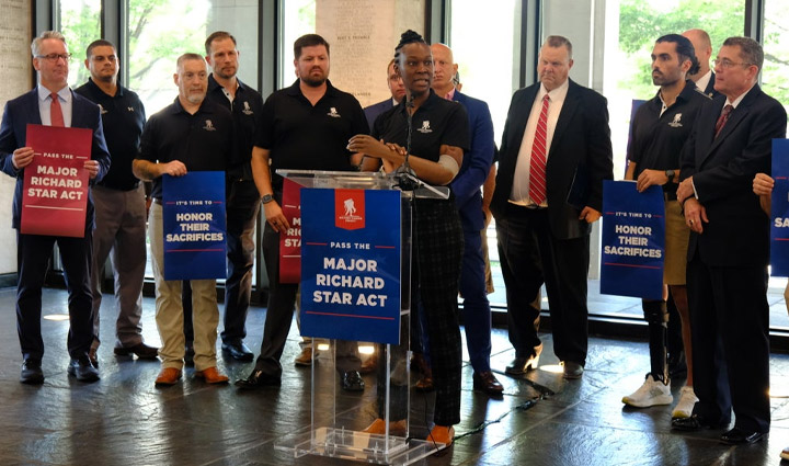 Wounded warrior Danielle Green stands at a podium with a microphone with a small crowd behind her while she speaks in support of the Major Richard Star Act.
