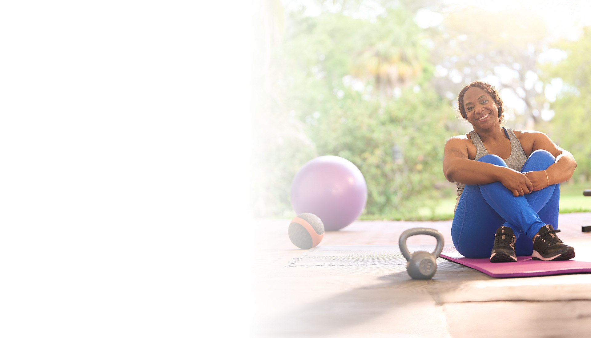 Wounded Warrior Yolanda Poulard in athletic clothing sits on an exercise mat outdoors, smiling and resting with her arms wrapped around her knees. Workout equipment is arranged around her.