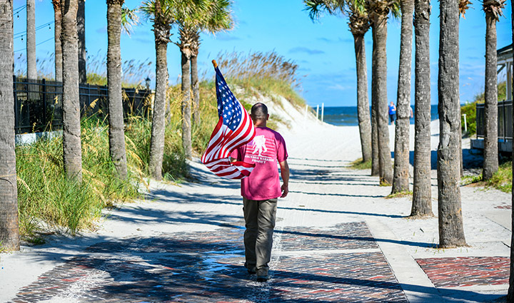 Wounded warrior James Rivera walking towards the beach on a sunny day wearing a pink WWP shirt and carrying an American flag.