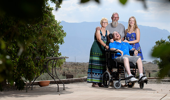 Wounded warrior Eric Schei is seated in his wheelchair smiling with his parents and sister standing behind him in an outdoor garden.